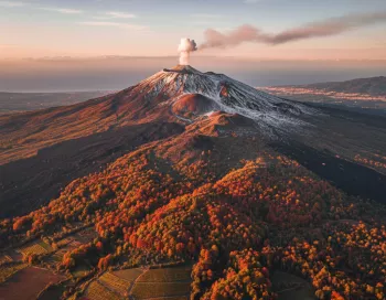 Vue aérienne du volcan Etna en automne avec des forêts aux couleurs orangées et les sommets enneigés sous un panache de fumée