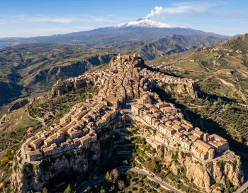 Vue aérienne par drone du village perché de Centuripe, révélant sa silhouette humaine unique étalée sur les crêtes rocheuses, avec l'Etna enneigé et fumant en arrière-plan